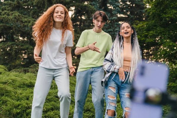 Energetic two girls and a boy dancing in front of a camera phone outdoors in the park. Friends recording video. Internet technology. People lifestyle.