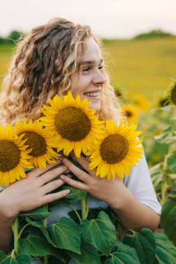 Genç kıvırcık saçlı kadın ayçiçeği tarlasında poz veriyor. Yaz ve seyahat kavramları.