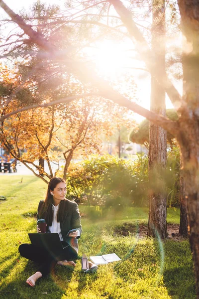 Barefoot young caucasian woman wearing jacket sitting on grass with laptop and single-use cup of coffee, searching information for her job. Young creative