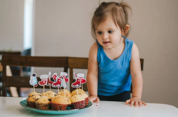The baby girl standing at the table and looking eagerly at the muffins on the plate that look so tasty being prepared for the Christmas table. Closeup for