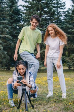 Yeşil parkta dans eden neşeli üç arkadaş, sosyal medyada paylaşmak için evdeki tripodla telefon kullanarak video çekiyorlar. İnsanların yaşam tarzı konsepti. İnternet
