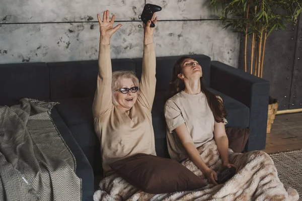 Caucasian senior woman celebrating holding hands up while playing video games with her daughter at home. Gaming and entertainment concept. Happy family. Family