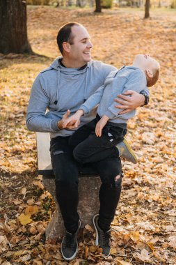 Beyaz bir baba, sonbaharda açık hava parkında eğlenirken oğlunu kollarında tutuyor. Mutlu aile, çocukluk. Sonbahar sezonu. Güneşli bir gün. Ebeveyn, çocuk. Eğlenceli.
