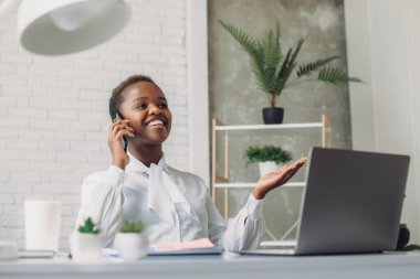 Modern ofiste laptopla çalışan mutlu siyahi kadın cep telefonundan arıyor. Uzaktan iletişim konsepti. Serbest, çevrimiçi eğitim veya sosyal medya kavramı