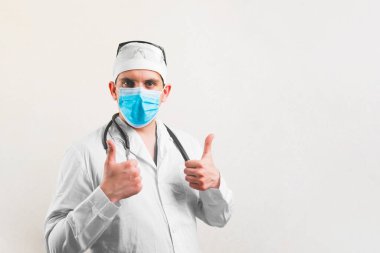 Beyaz takım elbiseli genç doktor portresi ve koruyucu solunum maskesi gözlüğü ve baş parmakları izole edilmiş steteskop.