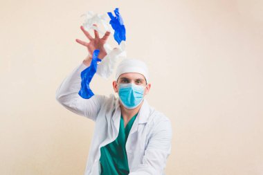 Beyaz önlüklü, koruyucu solunum maskeli genç doktor portresi sarı arka planda beyaz ve mavi eldivenler atıyor. Seçici odak.