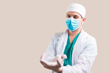 Beyaz takım elbiseli ve koruyucu maskeli genç doktor sarı arka planda eldivenlerini takıyor..
