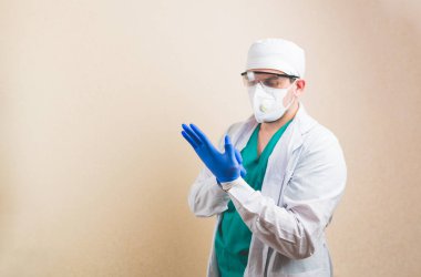 Beyaz takım elbiseli ve koruyucu maskeli genç doktor sarı arka planda eldivenlerini takıyor..