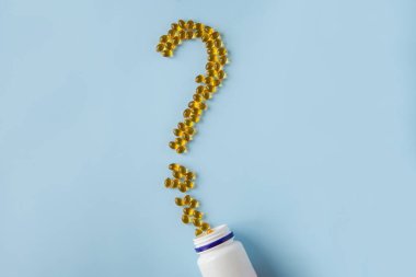 Mavi bir yüzey üzerinde soru işareti şeklinde vitamin kapsülleri. Omega ve D vitamini. Sağlık hizmeti. Besleyici takviyeler. Sarı haplar, haplar ve kapsüllerle açılmış bir şişe. İlaç soruları.