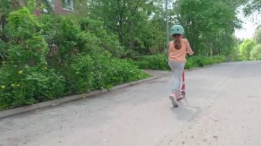The girl in the blue helmet rides a scooter along a European city street on a summer sunny day.