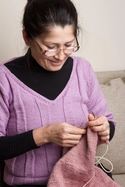 Asian woman knits on spokes a thing, sitting on an sofa in a room — Stock Photo, Image