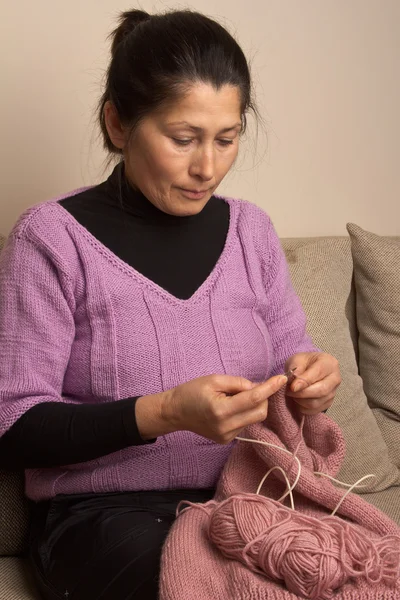 Asian woman knits on spokes a thing, sitting on an sofa in a room — Stock Photo, Image