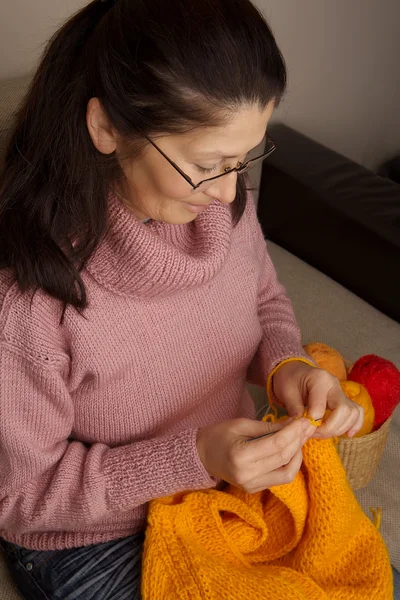 Mulher asiática tricota em spokes uma coisa, sentado em um sofá em um quarto — Fotografia de Stock