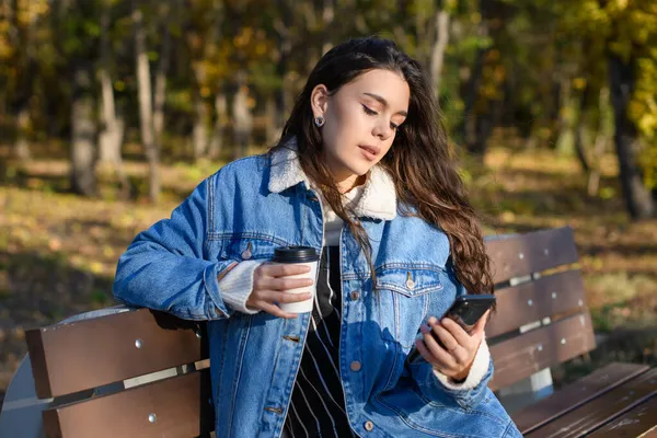 Молодая Женщина Скамейке Осеннем Парке Кофе Смартфоном Руках — стоковое фото