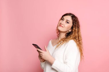 Elinde akıllı bir telefonla pembe bir arka planda duran neşeli ve çekici genç bir kadın kameraya bakar ve gülümser..