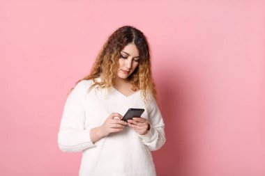 Pembe bir arka planda elinde akıllı bir telefonla yalnız duran neşeli, çekici bir genç kadın..