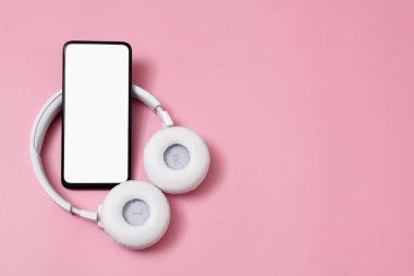 A smartphone with an empty white screen and wireless headphones on a pink pastel background. With a place for the inscription.
