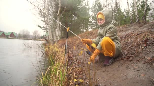 Kalın elbiseli bir çocuk gölün kıyısında balık tutuyor. Kız bir balık yakaladığı için mutlu. Çocuk şamandırayı işaret eder ve konuşur, gülümser.. — Stok video