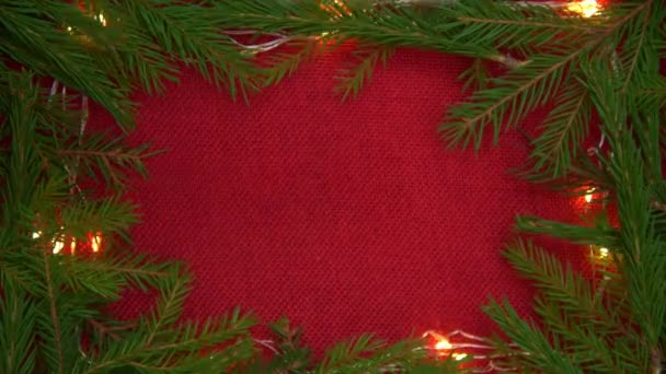 Предпосылки для Рождества. Красная ткань окружена еловыми ветвями и гирляндами. Рука кладет коробку с подарком в центр. Серебряная блестящая подарочная коробка, концепция поздравления с Новым годом и Рождеством — стоковое видео