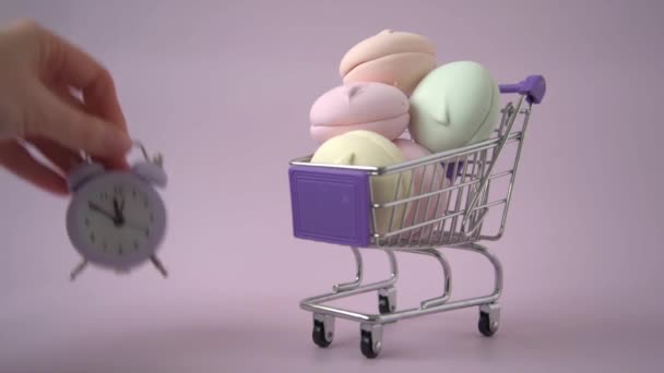 Nel carrello della spesa è un marshmallow colorato. Una mano mette una sveglia accanto ad essa. Il concetto di shelf life del prodotto. Il concetto di ricevere dolci in determinati momenti della giornata. — Video Stock