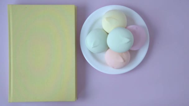 Zicht van bovenaf. Op tafel ligt een boek, een bord marshmallow. Een vrouwenhand voegt een mok cacao toe aan deze samenstelling. Gezellig en aangenaam plaatje in pastelkleuren. — Stockvideo