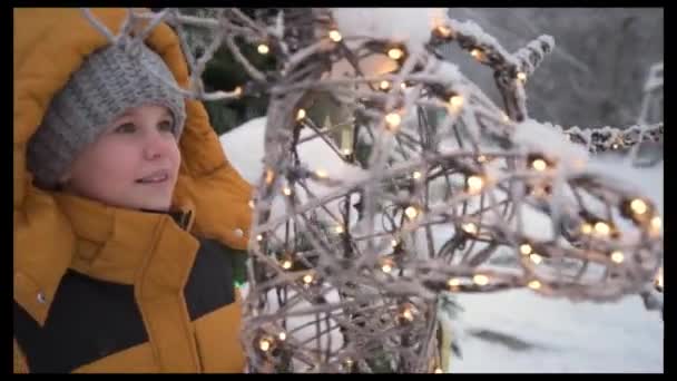 这孩子很羡慕地审视着小鹿的身影.一个穿着黄色夹克、头戴灰色针织帽子、衣着温暖的女孩走在白雪覆盖的街道上。每样东西都装饰成欢迎圣诞节 — 图库视频影像