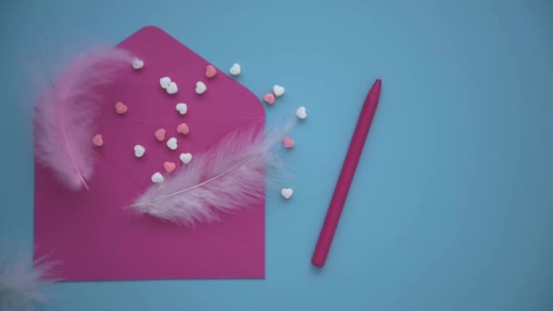 On a blue background lies a pink envelope. Hearts are scattered nearby, there is a pen. Delicate feathers complement the romantic image of the overall picture. The process of writing greetings for the — Stockvideo