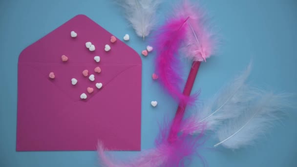 On a blue background lies a pink envelope. Hearts are scattered nearby, there is a pen. Delicate feathers complement the romantic image of the overall picture. The process of writing greetings for the — Stockvideo
