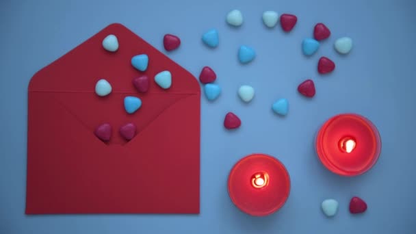 A stylish and cozy picture for Valentines Day. View from above, on a blue background laid out an envelope, candles and hearts. A red envelope from which hearts are poured. Burning candles create a — Stock Video