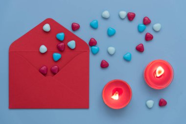 Sevgililer Günü için şık ve rahat bir fotoğraf. Yukarıdan bakıldığında, mavi arkaplanda bir zarf, mumlar ve kalpler vardı. Kalplerin döküldüğü kırmızı bir zarf. Yanan mumlar bir şey yaratır.