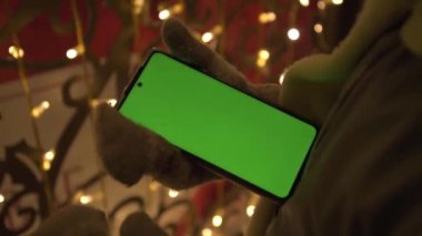 Eller eldiven içinde, Noel ışıklarının arka planına doğru. Sağ elinde yeşil ekranlı bir telefon var. Kromakey. Sol eliyle, çocuk telefondaki sayfaları çeviriyor.