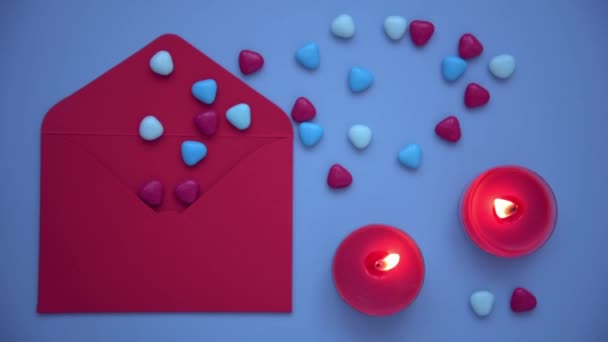 A stylish and cozy picture for Valentines Day. View from above, on a blue background laid out an envelope, candles and hearts. A red envelope from which hearts are poured. Burning candles create a — 图库视频影像