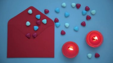 Sevgililer Günü için şık ve rahat bir fotoğraf. Yukarıdan bakıldığında, mavi arkaplanda bir zarf, mumlar ve kalpler vardı. Kalplerin döküldüğü kırmızı bir zarf. Yanan mumlar bir şey yaratır.