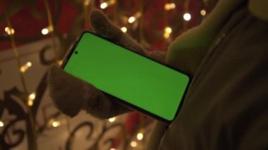 Eller eldiven içinde, Noel ışıklarının arka planına doğru. Sağ elinde yeşil ekranlı bir telefon var. Kromakey. Sol eliyle, çocuk telefondaki sayfaları çeviriyor.