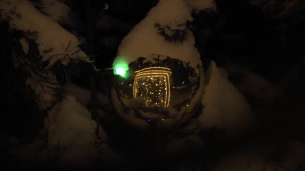 Les guirlandes se reflètent dans la grande boule de Noël. Vue de nuit. La boule de Noël, recouverte de neige, reflète la beauté autour. — Video