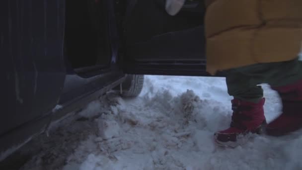 Το παιδί μπαίνει στο αυτοκίνητο και πριν αυτό χτυπήσει τις μπότες για να φύγει από το χιόνι. Μετά χτυπάει την πόρτα του αυτοκινήτου. Χιονισμένο χειμώνα. — Αρχείο Βίντεο