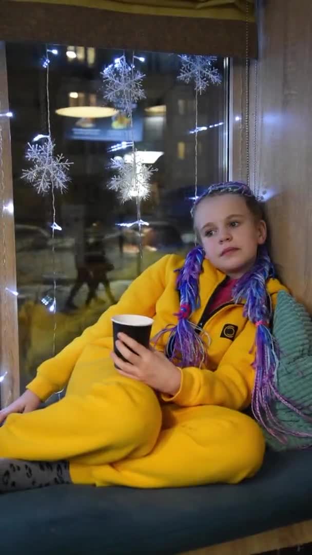 The teen age girl sat comfortably on the windowsill. The child pondered. The girl is sad. A beautiful girl with multi-colored braids in a yellow bright jumpsuit. He holds a glass of hot drink in his — ストック動画