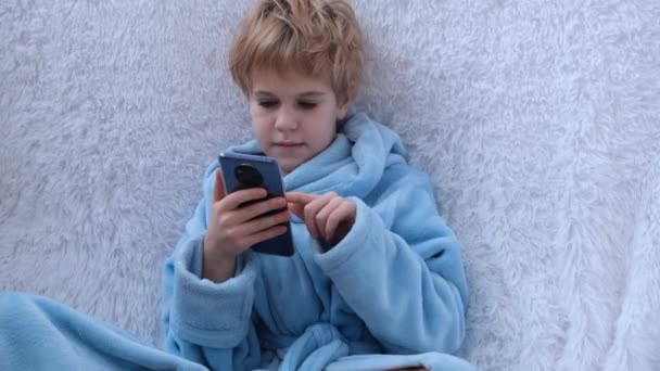 A menina sentou-se no sofá. Um adolescente em um roupão de banho acolhedor em um cobertor macio olha para um smartphone. Emoções, a menina sorri. — Vídeo de Stock