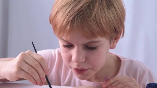 Close-up van het gezicht van de kindkunstenaars. Een kleine getalenteerde man schildert ijverig een schilderij met een kwast. Het gezicht drukt emoties uit. Focus en ernst. Mooi roodharig kind met een kort kapsel — Stockvideo