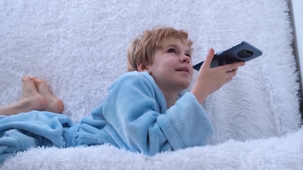 Uma adolescente dita uma mensagem de voz para um smartphone. Uma criança em um roupão de banho acolhedor no sofá fala em um smartphone. Ambiente acolhedor em casa. — Vídeo de Stock