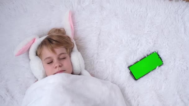 El concepto de felicitaciones por la Pascua. Hermosa adolescente duerme dulcemente. Bebé con orejas de conejo. Cerca se encuentra un teléfono con una pantalla verde. De repente el teléfono emite un pitido y el niño se despierta. A — Vídeos de Stock