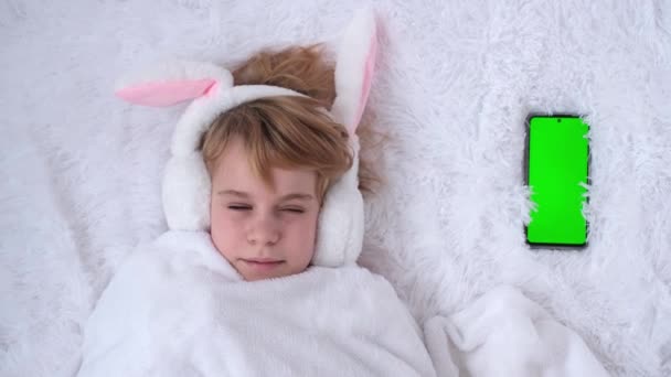 Koncept gratulace k Velikonocím. Krásná dospívající dívka sladce spí. Dítě s králičíma ušima. Nedaleko leží telefon se zelenou obrazovkou. Náhle pípne telefon a dítě se probudí. A — Stock video