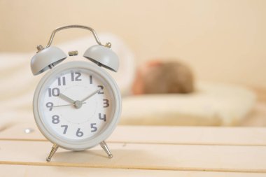 Gri alarm saati ahşap bir masanın üzerinde duruyor. Arka planda uyuyan bir adamla bir yatak var. Sabah konsepti, uyanış. Kopyalama alanı.
