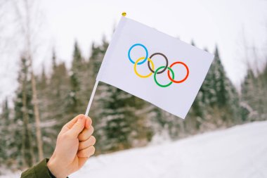 The Olympic flag, small in hand, flutters against the backdrop of snow and trees Concept for Winter Olympic Games 2022.	