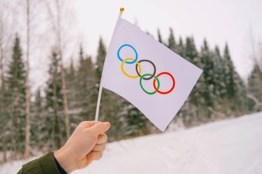 The Olympic flag, small in hand, flutters against the backdrop of snow and trees Concept for Winter Olympic Games 2022.	