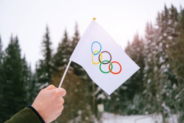 The Olympic flag, small in hand, flutters against the backdrop of snow and trees Concept for Winter Olympic Games 2022.	