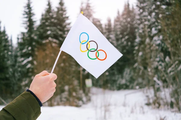 The Olympic flag, small in hand, flutters against the backdrop of snow and trees Concept for Winter Olympic Games 2022.	