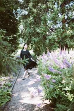 Beautiful happy middle-aged Japanese woman in black casual dress and wedge heel flip flops summer park, smiling. Sunlight, copy space