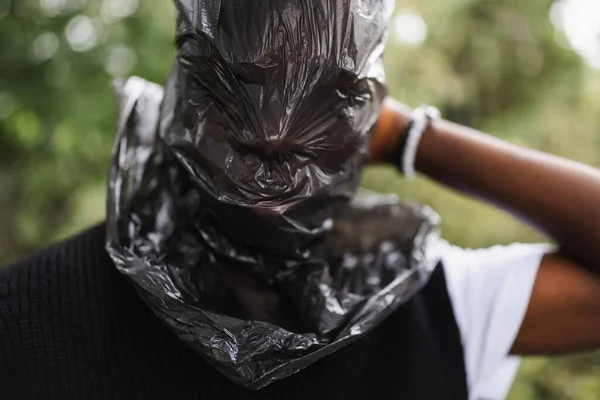 Young African man outdoor with trash bag over his head, street portrait. Plastic pollution, ecology, environmental conversation, mental health concept. Excess of plastic in nature and life