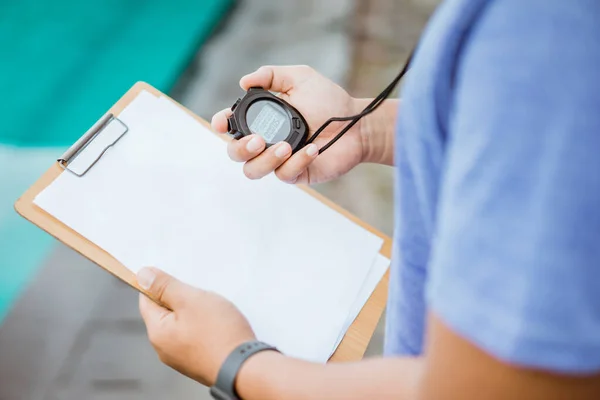 Close up of a coachs hand showing time with stopwatch and clipbord for notes while standing — Stock Photo, Image